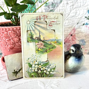 May include: Vintage Easter postcard with the text "A Peaceful Easter" in ornate lettering. The postcard features a cross-shaped design with a landscape scene of a church, road, and flowers. A ceramic bird figurine and a pink flower pot are also in the image.