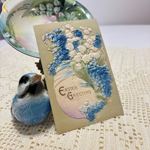 Postal antigua de felicitación de Pascua de 1911: nomeolvides azules en bajorrelieve y lirios del valle con huevo de Pascua blanco y lavanda en relieve