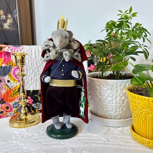 Puede incluir: Una figura caprichosa de un rey ratón, con una corona dorada, una capa roja y una chaqueta azul con adornos dorados. El rey ratón tiene múltiples cabezas de ratón alrededor del cuello y sostiene una batuta de director. La figura se encuentra sobre una base verde.