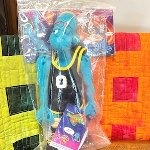 May include: A blue plush figure of a Space Jam character, wearing a black and yellow basketball jersey with the number 0. The figure is in a clear plastic bag with a tag that reads "Warner Bros." and "Space Jam". The figure has black and white sneakers.