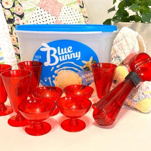 Puede incluir: Una colección de cristalería roja, que incluye copas, cuencos y vasos altos, está dispuesta cerca de un recipiente de helado Blue Bunny. También es visible una cuchara para helado roja. La escena sugiere un ambiente temático de postre.
