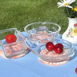 Peut inclure: Nature morte avec des bols en verre transparent et un petit vase fleuri. Les bols contiennent des fruits frais, dont des fraises et des cerises, posés sur des dessous de verre en crochet. La scène se déroule sur une nappe bleue.