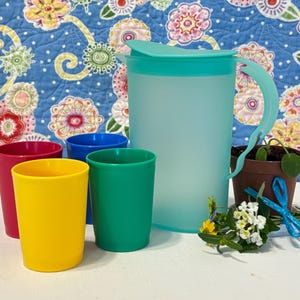 May include: A collection of colorful plastic drinkware. Includes a large turquoise pitcher with a matching lid and handle, and five cups in red, yellow, blue, and green. A small potted plant and flowers are also present.