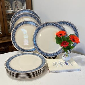 May include: A set of cream-colored dinner plates with a blue floral border. The plates are stacked and arranged on a white tablecloth, with a small vase of orange flowers and a book titled "How to Make an American Quilt".