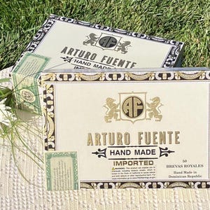 May include: Two wooden cigar boxes with gold lettering and a gold crest. The top box says "ARTURO FUENTE HAND MADE IMPORTED" and the bottom box says "ARTURO FUENTE HAND MADE IMPORTED 50 BREVAS ROYALES Hand Made in Dominican Republic".