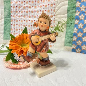 Puede incluir: Una figurita de porcelana de una niña con el pelo rojo, que lleva un vestido rojo con lunares blancos, tocando una guitarra pequeña. Está de pie sobre una base blanca.