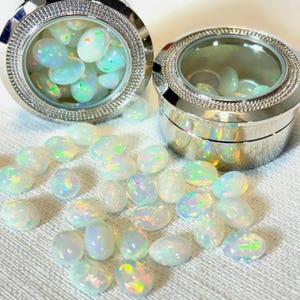May include: Oval-shaped opals with iridescent color play are scattered on a white surface. Two silver containers with clear lids are filled with similar opals. The opals display flashes of green, blue, and yellow.