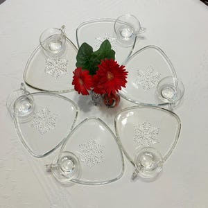 May include: Set of six clear glass triangular plates with snowflake designs. Each plate has a matching glass cup with a handle. A bouquet of red flowers is in the center of the plates.