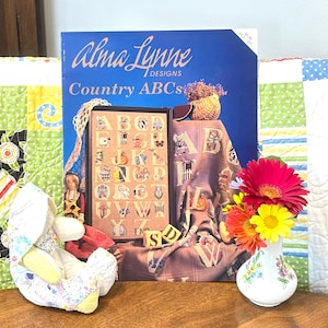 May include: A still life featuring a cross-stitch pattern book titled "Alma Lynne Designs Country ABCs" with a framed sampler. A stuffed bunny, a small vase with flowers, and a quilt are also present. The book is blue with white text.
