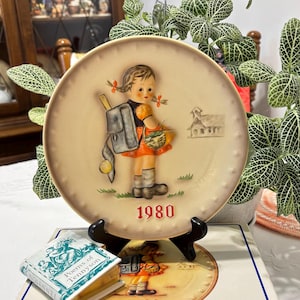 May include: A vintage 1980 collectible plate featuring a girl with a backpack and a basket of apples walking towards a schoolhouse. The plate is cream-colored with a red and black border.