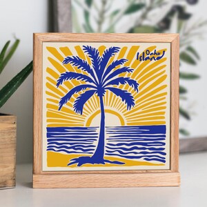 May include: A wooden framed square sign with a yellow background and a blue palm tree silhouette. The sun is setting behind the palm tree and the words 'Oahu Island' are in the top right corner.
