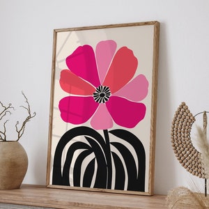 Op de afbeelding: Een ingelijste kunstdruk met een grote, gestileerde bloem in roze en rode tinten op een cr&egrave;mekleurige achtergrond. De bloem heeft een zwart hart en staat op een zwarte, gebogen stengel. Het kunstwerk wordt tentoongesteld op een houten kast.