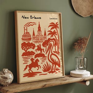 Op de afbeelding: Een ingelijste kunstprint met een rode illustratie van New Orleans, met een pelikaan, palmbomen en architecturale elementen. De woorden "New Orleans" en "Louisiana" staan bovenaan.