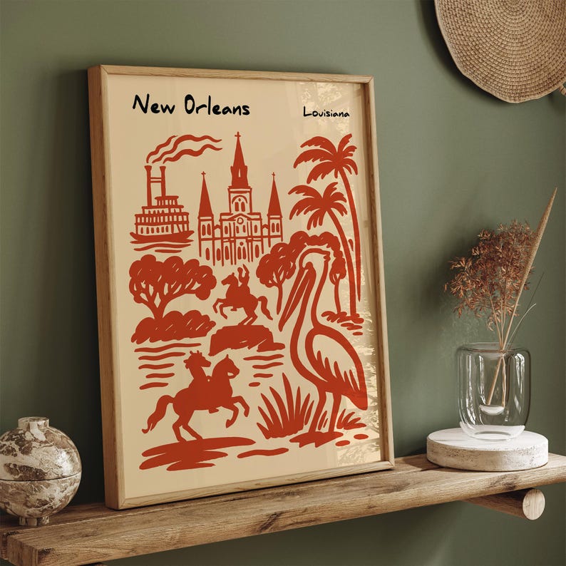 Op de afbeelding: Een ingelijste kunstprint met een rode illustratie van New Orleans, met een pelikaan, palmbomen en architecturale elementen. De woorden "New Orleans" en "Louisiana" staan bovenaan.