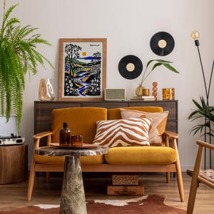 May include: A mustard yellow velvet sofa with a zebra print throw pillow and a framed print of a landscape with the word "Vermont" on it. The sofa is in a living room with a wooden coffee table, a record player, and a potted plant.