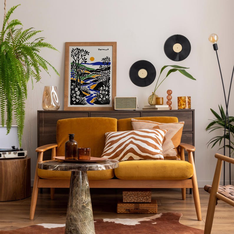 May include: A mustard yellow velvet sofa with a zebra print throw pillow and a framed print of a landscape with the word "Vermont" on it. The sofa is in a living room with a wooden coffee table, a record player, and a potted plant.