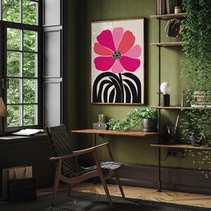 Op de afbeelding: Een ingelijste kunstafdruk met een grote, gestileerde bloem in roze en rode tinten, met een zwart centrum en steel. Het kunstwerk hangt in een kamer met een houten bureau, stoel en planken, wat een modern interieur cre&euml;ert.