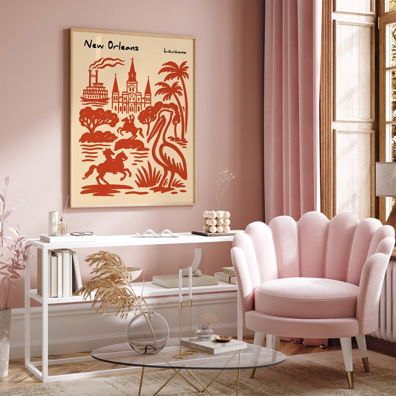 Op de afbeelding: Een ingelijste kunstprint met een rood-beige illustratie van bezienswaardigheden in New Orleans, waaronder een raderstoomboot, kathedraal en pelikaan. De tekst "New Orleans Louisiana" staat bovenaan. De print wordt getoond in een roze kamer.