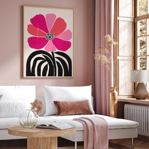 Op de afbeelding: Een ingelijste kunstdruk met een grote, gestileerde bloem in roze en rode tinten op een cr&egrave;mekleurige achtergrond. De stengel en basis van de bloem zijn zwart. Het kunstwerk wordt tentoongesteld in een woonkamer met een bank en decoratieve elementen.