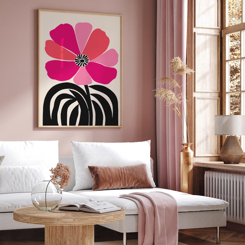 Op de afbeelding: Een ingelijste kunstdruk met een grote, gestileerde bloem in roze en rode tinten op een cr&egrave;mekleurige achtergrond. De stengel en basis van de bloem zijn zwart. Het kunstwerk wordt tentoongesteld in een woonkamer met een bank en decoratieve elementen.