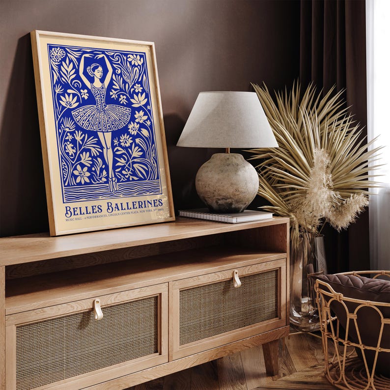 May include: A blue and white poster with a stylized illustration of a ballerina dancing in a field of flowers. The poster is framed in a light wood frame and is titled "Belles Ballerines".