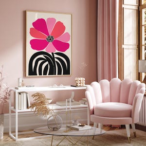 Op de afbeelding: Een ingelijste kunstprint met een grote, gestileerde bloem in roze en rode tinten op een cr&egrave;mekleurige achtergrond. De bloem staat boven zwarte, gebogen lijnen. De print hangt in een roze kamer met een roze stoel.