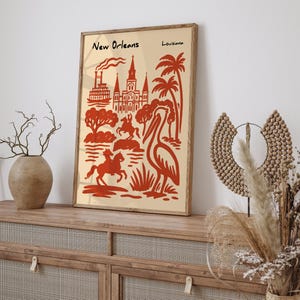 Op de afbeelding: Een ingelijste kunstprint met een rode en beige illustratie van New Orleans, Louisiana. Het kunstwerk toont bezienswaardigheden, palmbomen en een pelikaan. De woorden "New Orleans" en "Louisiana" staan bovenaan de print.