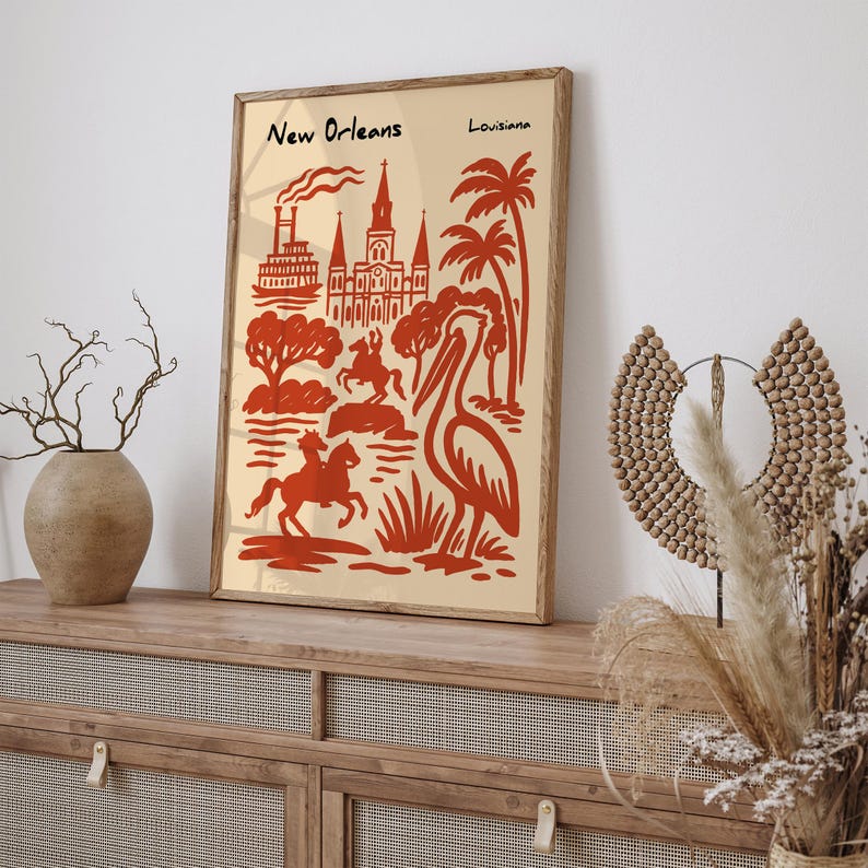 Op de afbeelding: Een ingelijste kunstprint met een rode en beige illustratie van New Orleans, Louisiana. Het kunstwerk toont bezienswaardigheden, palmbomen en een pelikaan. De woorden "New Orleans" en "Louisiana" staan bovenaan de print.