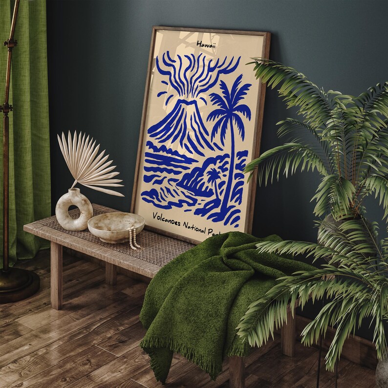 Puede incluir: Una impresi&oacute;n art&iacute;stica enmarcada con una ilustraci&oacute;n azul de un volc&aacute;n, una palmera y una escena oce&aacute;nica, con el texto "Hawaii" y "Volcanoes National Park". La impresi&oacute;n se exhibe en un banco de madera con una manta verde y objetos decorativos.