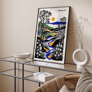 May include: A black framed print with a stylized landscape scene in blue, green, yellow, and white. The print features a winding river, trees, and flowers. The text "Vermont" is at the top of the print.