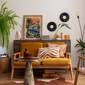 May include: A cozy living room scene with a mustard yellow sofa, a marble-topped coffee table, and a wooden cabinet. The room is decorated with a framed print, vinyl records, and decorative pillows. A fern and a floor lamp add to the ambiance.