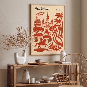 Op de afbeelding: Een ingelijste kunstdruk met een rode illustratie van bezienswaardigheden van New Orleans, waaronder een kathedraal, palmbomen en een pelikaan, tegen een cr&egrave;mekleurige achtergrond. De woorden "New Orleans" en "Louisiana" staan bovenaan.