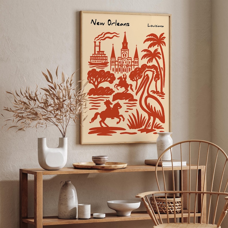 Op de afbeelding: Een ingelijste kunstdruk met een rode illustratie van bezienswaardigheden van New Orleans, waaronder een kathedraal, palmbomen en een pelikaan, tegen een cr&egrave;mekleurige achtergrond. De woorden "New Orleans" en "Louisiana" staan bovenaan.