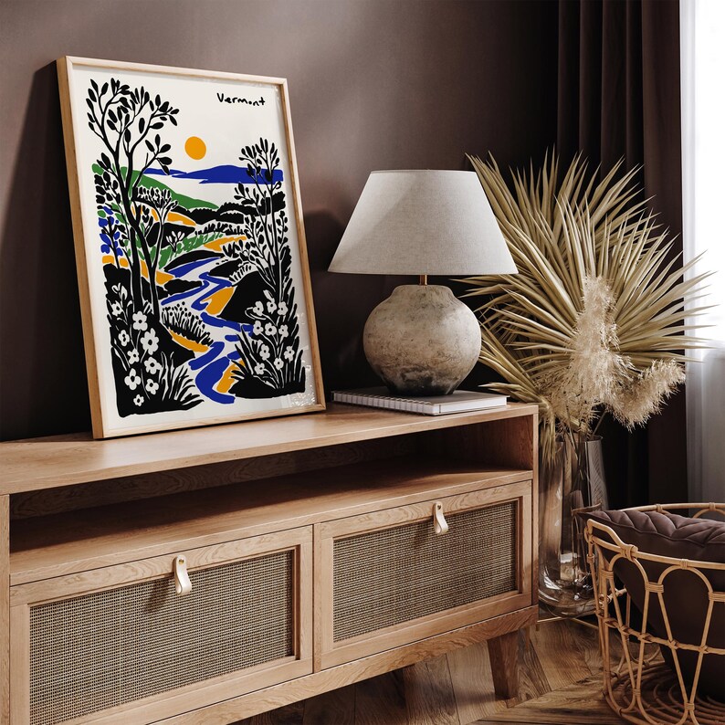 May include: A framed illustration of a landscape with a river, trees, and flowers. The illustration is in a black, white, blue, and yellow color scheme. The frame is made of wood and has a light brown finish. The illustration is titled "Vermont".