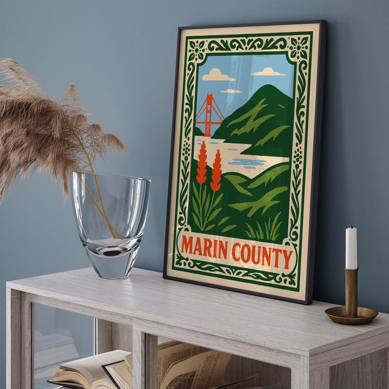 May include: A framed poster of Marin County, California, with a stylized illustration of the Golden Gate Bridge, green hills, and blue water. The poster has a decorative green border and the words "Marin County" in red. A clear vase and a candle are on a light-coloured wooden table.