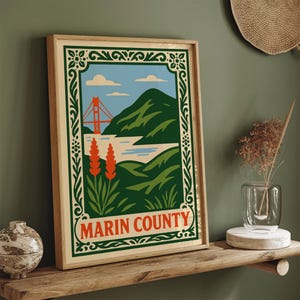 May include: Framed art print with a stylized illustration of Marin County. The artwork shows a red bridge, green hills, blue sky, and the text "Marin County" in orange. The print is in a wooden frame.