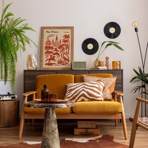Op de afbeelding: Een gezellige woonkamer met een mosterdgele bank, kussens met zebra- en streepmotief en een salontafel met marmeren blad. Een ingelijste New Orleans-print en twee vinylplaten sieren de muur. Een varen en een staande lamp zorgen voor groen.