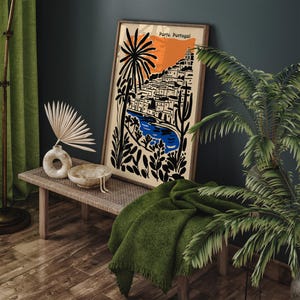 May include: A framed art print featuring a stylized illustration of Porto, Portugal, with a vibrant orange sky, blue river, and black and white buildings. The print is displayed on a wooden bench with a green blanket and decorative items.