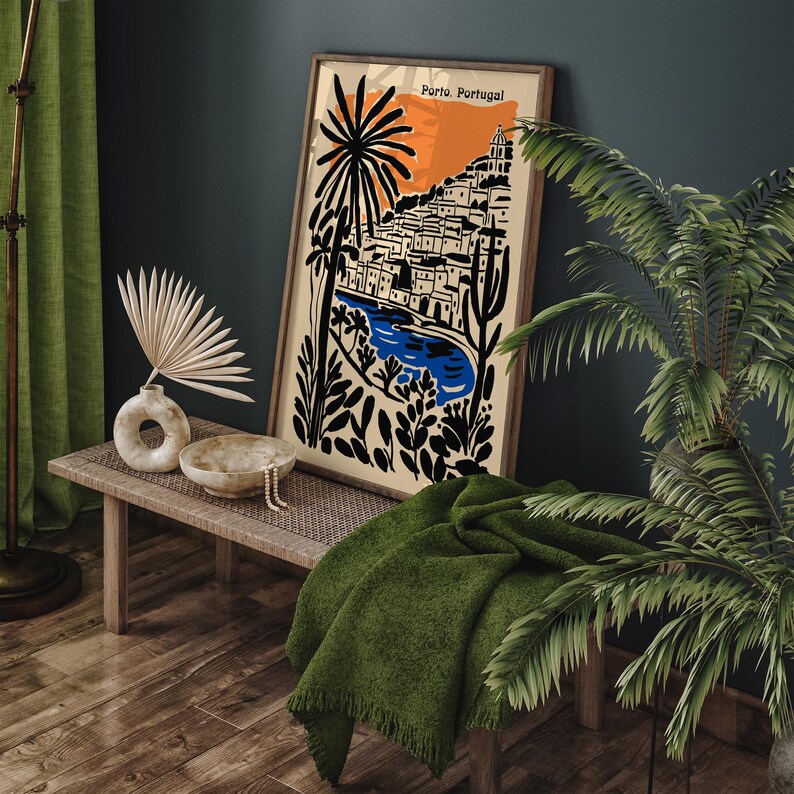 May include: A framed art print featuring a stylized illustration of Porto, Portugal, with a vibrant orange sky, blue river, and black and white buildings. The print is displayed on a wooden bench with a green blanket and decorative items.