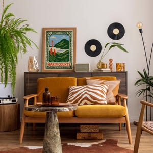 May include: A stylish living room featuring a mustard yellow sofa, a marble-topped side table, and a framed "Marin County" poster. Decorative cushions, a wooden cabinet, and vinyl records contribute to the retro style. A fern and a floor lamp complete the decor.