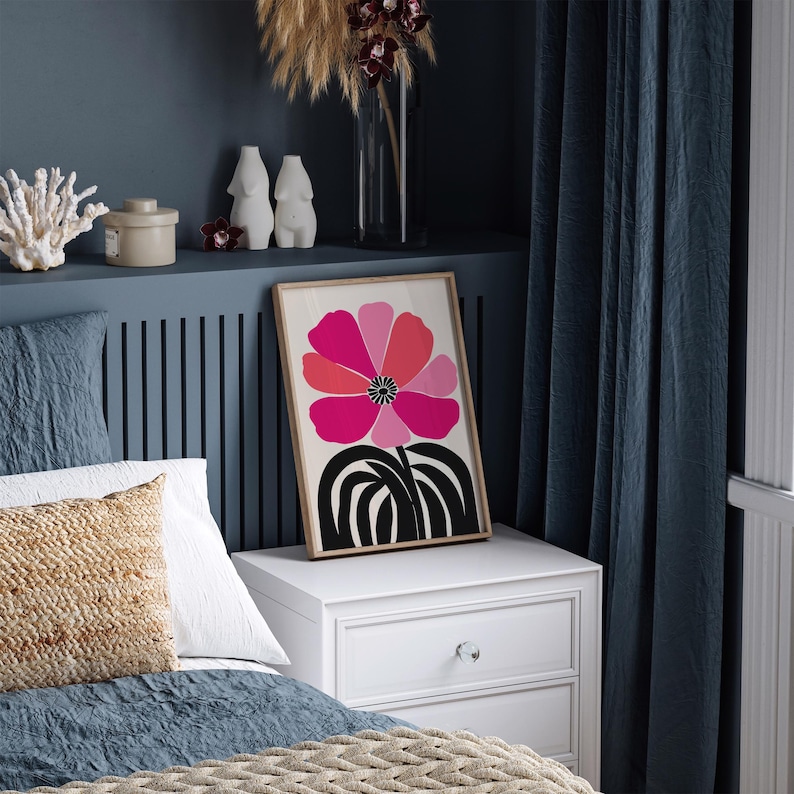 Op de afbeelding: Een ingelijste kunstprint met een gestileerde bloem in roze en rode tinten, met een zwart-witte basis. Het kunstwerk staat op een witte nachtkastje in een slaapkamer met blauwe muren en gordijnen.