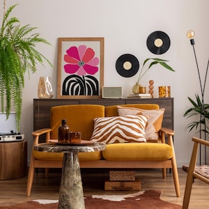 Op de afbeelding: Een modern woonkamer met een mosterdgele bank, een bijzettafel met marmeren blad en een ingelijste bloemenprint. Decoratieve elementen zijn vinylplaten, een luidspreker en diverse vazen. Een varen en een vloerlamp voegen groen en licht toe.