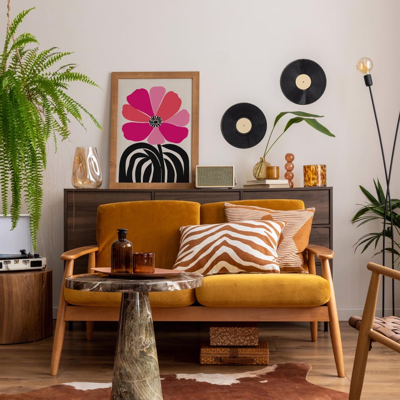Op de afbeelding: Een modern woonkamer met een mosterdgele bank, een bijzettafel met marmeren blad en een ingelijste bloemenprint. Decoratieve elementen zijn vinylplaten, een luidspreker en diverse vazen. Een varen en een vloerlamp voegen groen en licht toe.