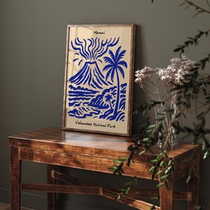 Puede incluir: Un estampado art&iacute;stico enmarcado con una ilustraci&oacute;n azul de un volc&aacute;n, palmeras y olas del oc&eacute;ano sobre un fondo beige. El estampado est&aacute; etiquetado como "Hawaii" y "Volcanoes National Park". El marco es de color marr&oacute;n oscuro.