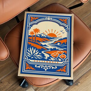 May include: A rectangular framed print featuring a stylized California landscape illustration in blue, orange, and cream. The artwork depicts palm trees, mountains, a sun, and flowers with the text CALIFORNIA printed at the bottom center.