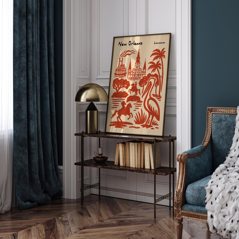 Op de afbeelding: Een ingelijste kunstdruk met een rode en beige illustratie van bezienswaardigheden en symbolen van New Orleans. De print bevat de tekst "New Orleans" en "Louisiana". Het kunstwerk wordt tentoongesteld op een donkerbruin rek met een gouden lamp.