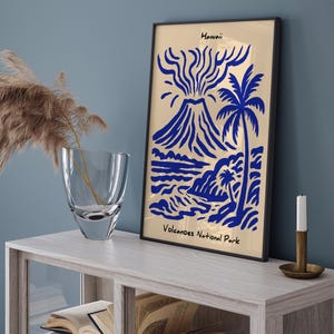 Puede incluir: Una impresi&oacute;n art&iacute;stica enmarcada con una ilustraci&oacute;n azul y dorada de un volc&aacute;n, una palmera y una escena oce&aacute;nica. La impresi&oacute;n est&aacute; etiquetada como "Hawaii" y "Volcanoes National Park". La obra de arte se exhibe sobre una mesa de madera clara.