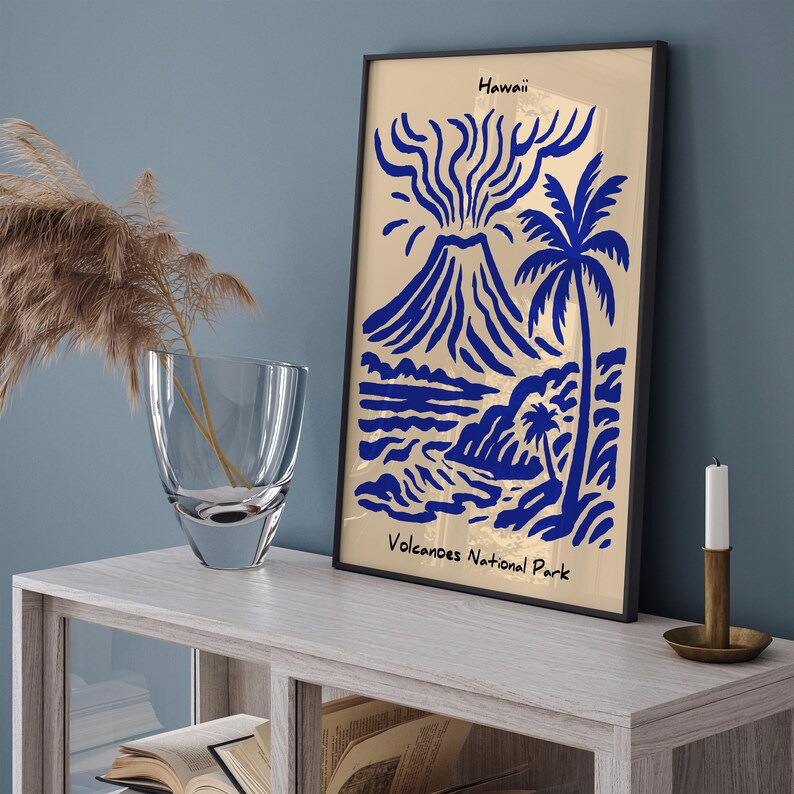 Puede incluir: Una impresi&oacute;n art&iacute;stica enmarcada con una ilustraci&oacute;n azul y dorada de un volc&aacute;n, una palmera y una escena oce&aacute;nica. La impresi&oacute;n est&aacute; etiquetada como "Hawaii" y "Volcanoes National Park". La obra de arte se exhibe sobre una mesa de madera clara.