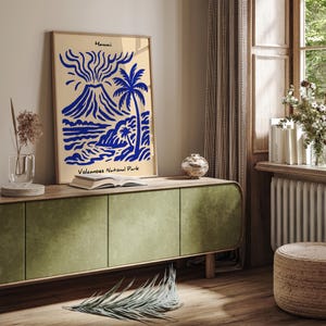 Puede incluir: Una impresi&oacute;n art&iacute;stica enmarcada con una ilustraci&oacute;n azul y beige de un volc&aacute;n, una palmera y una escena costera. El texto "Hawaii" y "Volcanoes National Park" es visible. La impresi&oacute;n se muestra en un mueble verde.