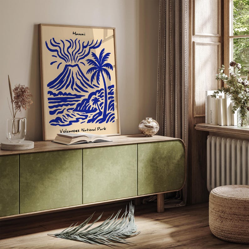 Puede incluir: Una impresi&oacute;n art&iacute;stica enmarcada con una ilustraci&oacute;n azul y beige de un volc&aacute;n, una palmera y una escena costera. El texto "Hawaii" y "Volcanoes National Park" es visible. La impresi&oacute;n se muestra en un mueble verde.